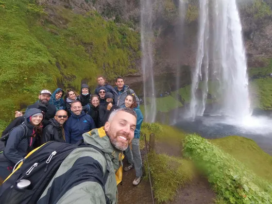Eine lachende WeRoad-Gruppe macht ein Selfie auf einem Weg neben einem großen Wasserfall, der sich eine grüne, moosige Felswand hinabstürzt.