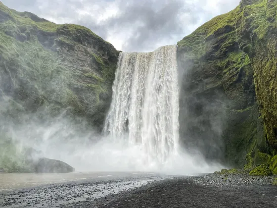 Ein breiter Wasserfall stürzt über eine moosgrüne Felswand und sprüht über einen schwarzen Kieselstrand unter einem grauen, bewölkten Himmel.