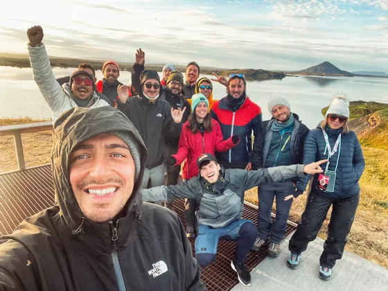 Ein Mann macht ein Selfie mit einer WeRoad-Gruppenreise an einem Aussichtspunkt mit Blick auf einen See und Hügel unter einem teilweise bewölkten Himmel.