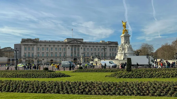 A grand classical palace and a golden-topped memorial statue stand behind a lawn and a road with people and traffic.