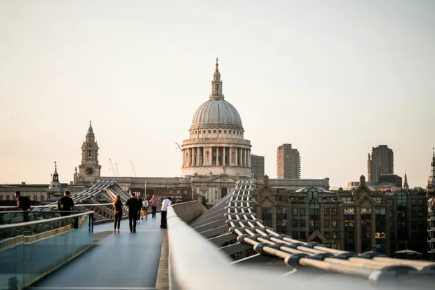 People walk on a modern pedestrian bridge towards a large, historic domed cathedral against a pale sky at sunset.