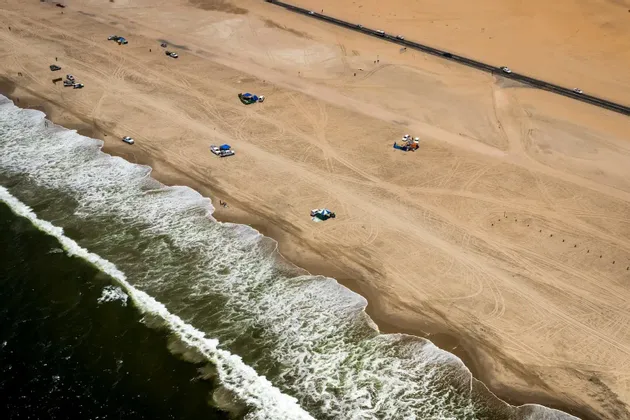 Une vue aérienne d'une plage de sable où l'océan rencontre le rivage, avec des véhicules et des tentes sur le sable et une route parallèle à la côte.
