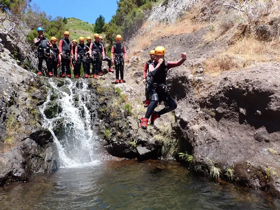 Eine WeRoad-Gruppenreise in Neoprenanzügen und Helmen beobachtet, wie eine Person von einem Felsen in einen natürlichen Pool an einem Wasserfall springt.