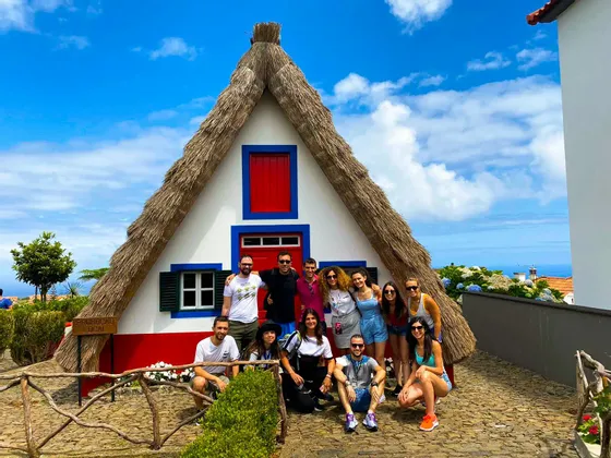 Eine WeRoad-Gruppenreise posiert an einem sonnigen Tag vor einem traditionellen Reetdachhaus mit A-Rahmen für ein Foto.