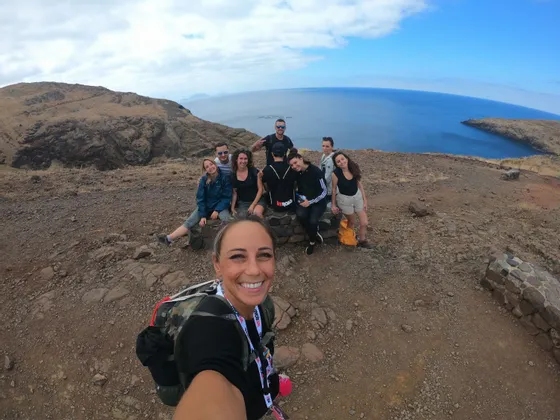 Eine lächelnde Frau macht ein Selfie mit ihrer WeRoad-Gruppenreise an einer felsigen Klippe mit Blick auf das Meer unter einem bewölkten blauen Himmel.