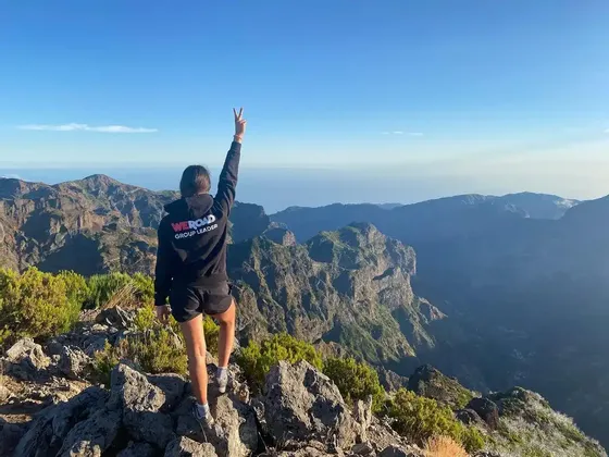 Ein WeRoad-Gruppenleiter steht mit dem Rücken zur Kamera auf einem felsigen Berggipfel und macht ein Friedenszeichen über einer weiten Bergkette.