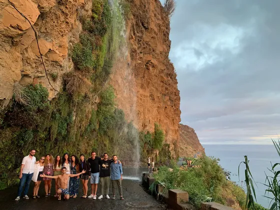 Eine WeRoad Gruppenreise posiert auf einer Küstenstraße unter einem Wasserfall, der von einer felsigen Klippe herabstürzt.