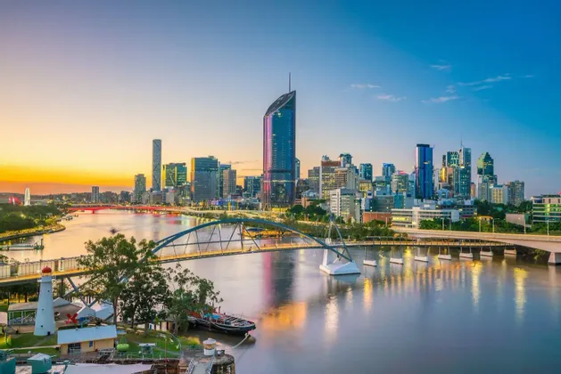 Moderne Skyline mit beleuchteten Wolkenkratzern, die sich bei Sonnenuntergang in einem breiten Fluss spiegeln, mit einer Fußgängerbrücke im Vordergrund.