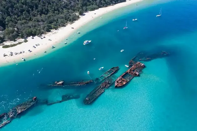 Ein Luftbild von mehreren versunkenen Schiffswracks, die in seichtem, türkisfarbenem Wasser an einem sandigen, von Bäumen gesäumten Ufer mit Booten liegen.