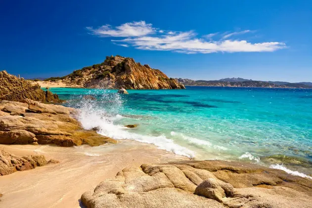 Un'onda si infrange contro grandi rocce su una spiaggia sabbiosa con acqua limpida e turchese sotto un cielo azzurro brillante con poche nuvole.