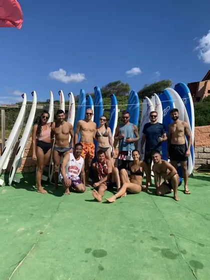 Un gruppo WeRoad in costume da bagno posa per una foto davanti a una rastrelliera di tavole da surf in una giornata di sole.