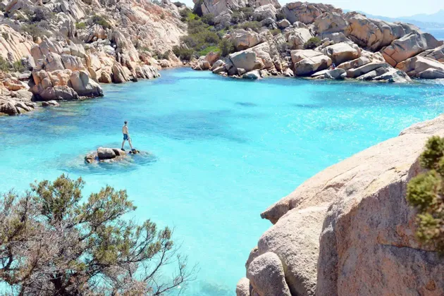 Una persona in costume da bagno si trova su una piccola roccia in una caletta con acqua cristallina turchese, circondata da scogliere rocciose.