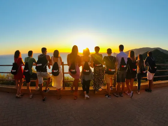 Un viaggio di gruppo WeRoad visto da dietro, mentre si osserva un tramonto dorato sul mare da un punto panoramico costiero.