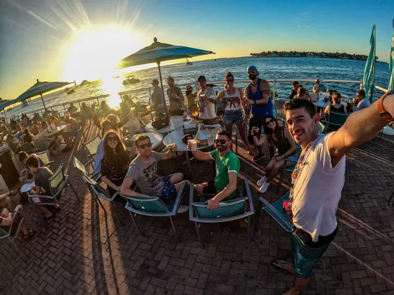Un gruppo WeRoad scatta un selfie su una terrazza sul mare al tramonto, con bevande sui tavoli.