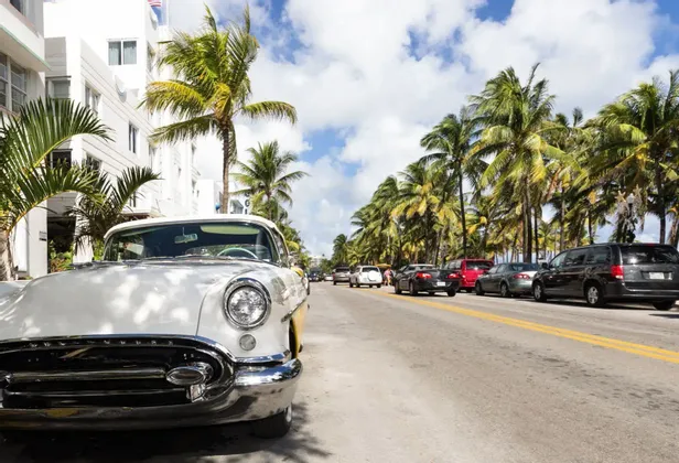 Un'auto d'epoca bianca parcheggiata su una strada assolata fiancheggiata da alte palme accanto a un edificio bianco.