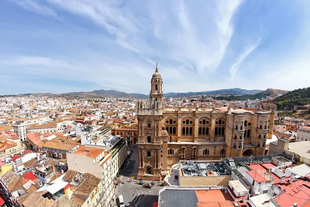 Una vista aerea di una cattedrale storica circondata da un paesaggio urbano con tetti di tegole rosse e montagne sullo sfondo.