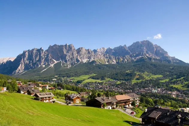 Un villaggio alpino con edifici in legno è immerso in una valle verde ai piedi di una catena montuosa rocciosa.