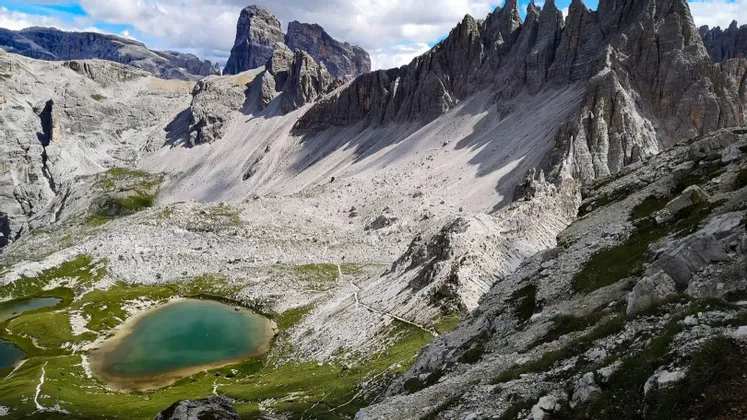 Vista dall'alto di un lago alpino turchese in una vasta valle rocciosa con cime frastagliate sotto un cielo parzialmente nuvoloso.