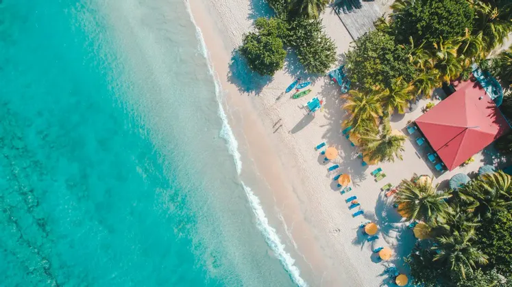 Panoramica aerea di una spiaggia tropicale con acqua turchese, sabbia bianca, palme, kayak colorati e un edificio dal tetto rosso.