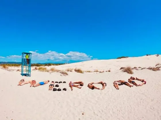 Un viaje en grupo de WeRoad tumbados en una playa de arena blanca, usando sus cuerpos y mochilas para deletrear la palabra WEROAD bajo un cielo azul.