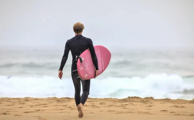 Un surfista in muta nera, visto di spalle, porta una tavola da surf rosa mentre cammina su una spiaggia sabbiosa verso l'oceano.