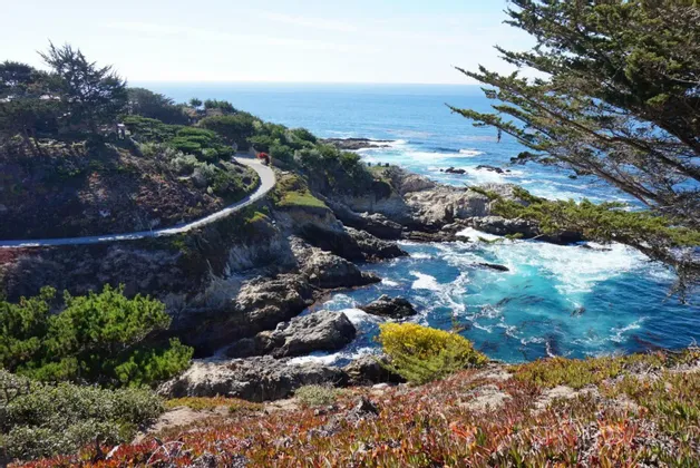 Una vista dall'alto di una strada tortuosa lungo una costa rocciosa con acqua turchese e vegetazione verde sotto un cielo sereno.