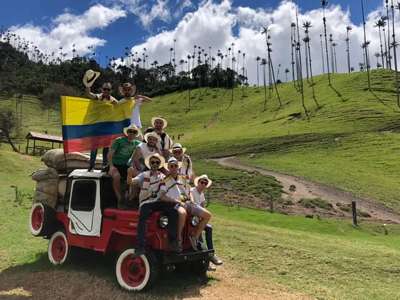 Un viaje en grupo de WeRoad posa en un jeep rojo, sosteniendo una bandera colombiana, en un valle verde con palmeras altas.