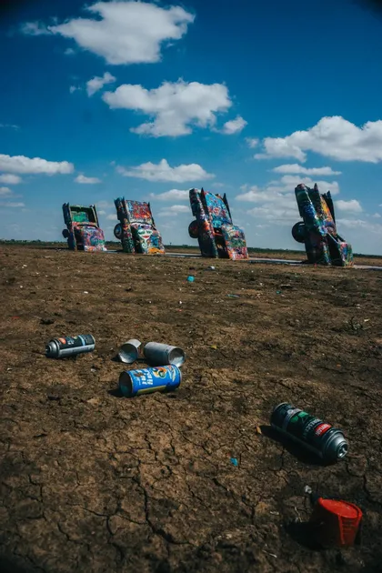 Mit Graffiti bedeckte Autos, kopfüber auf einem Feld vergraben, mit leeren Sprühdosen auf der rissigen Erde unter einem blauen, bewölkten Himmel.