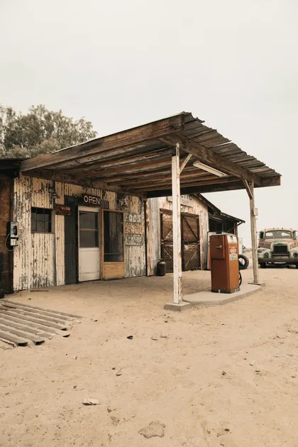 Eine rustikale Tankstelle mit Wellblechwänden, einer alten orangen Zapfsäule und einem alten LKW, der auf einem sandigen Parkplatz steht.