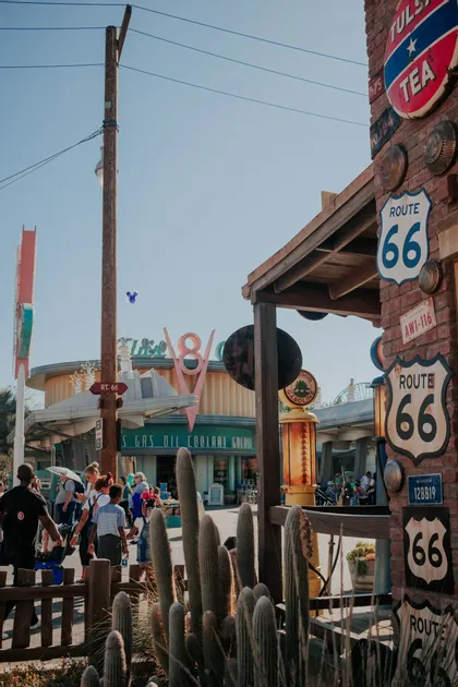 Eine sonnige Straßenszene mit Route 66-Schildern an einem Backsteingebäude, Menschen, die an Kakteen vorbeigehen, und einem Retro-V8-Schild im Hintergrund.
