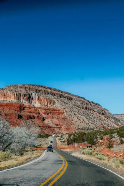 Automobili percorrono una strada tortuosa accanto a una grande montagna di roccia rossa stratificata sotto un cielo azzurro e luminoso.