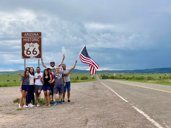 Un gruppo WeRoad in viaggio posa per una foto accanto a un cartello della storica Route 66 in Arizona, con una persona che sventola una bandiera americana.