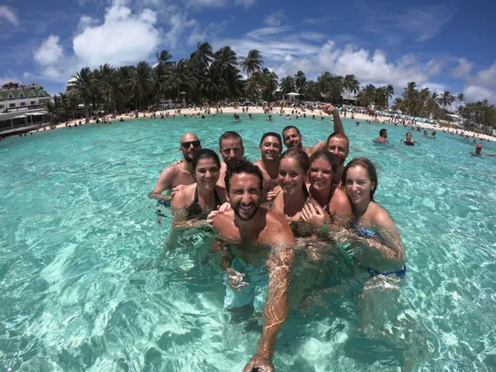 Un grupo de WeRoad se toma una selfie sonriendo en las aguas claras y turquesas del océano, con una playa con palmeras y muy concurrida de fondo.