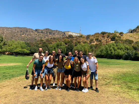 Eine WeRoad-Gruppenreise posiert für ein Foto in einem grasbewachsenen Park, mit dem Hollywood-Schriftzug auf einem Hügel im Hintergrund unter einem klaren Himmel.