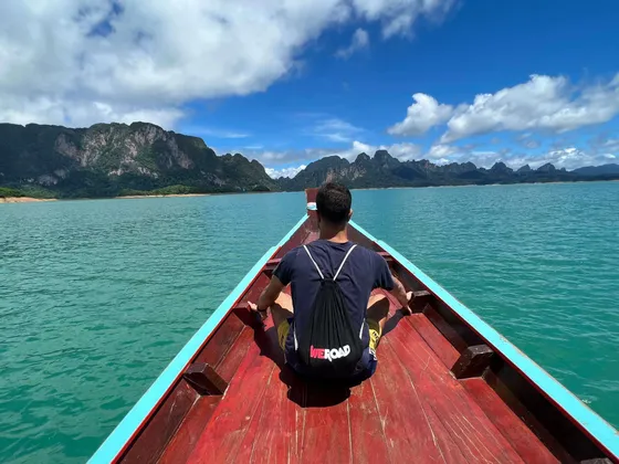 Une personne portant un sac à dos WeRoad est assise à la proue d'un bateau en bois sur un lac turquoise, avec des montagnes vertes au loin.