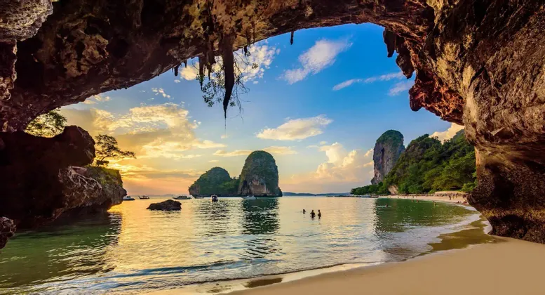 Vue depuis l'intérieur d'une grotte marine surplombant une plage tropicale avec des karsts calcaires et des bateaux au coucher du soleil.