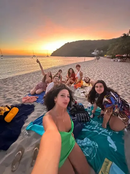 Une femme prend un selfie de son groupe WeRoad lors d'un pique-nique sur une plage de sable, au coucher du soleil coloré.