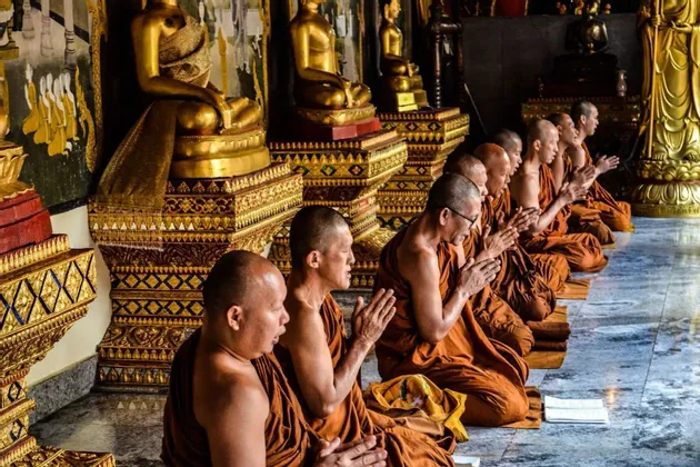 Eine Reihe buddhistischer Mönche in orangefarbenen Roben kniet betend in einem Tempel, mit goldenen Buddha-Statuen im Hintergrund.