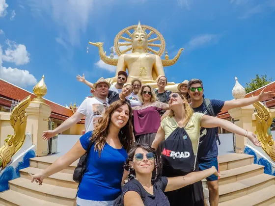 Eine WeRoad-Gruppenreise lächelt und posiert für ein Foto auf einer Treppe vor einer großen goldenen Buddha-Statue unter blauem Himmel.