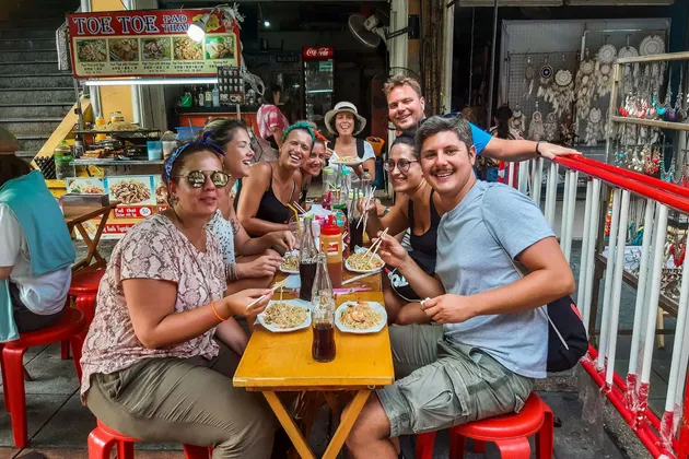 Un grupo de WeRoad sonriente come fideos con palillos en una mesa al aire libre frente a un puesto de comida callejera.