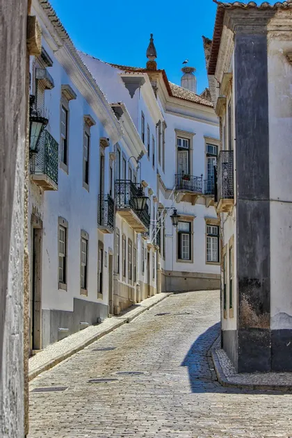 Una stretta strada acciottolata e assolata si inerpica tra edifici imbiancati a calce, con un nido di cicogna su un tetto contro un cielo azzurro.
