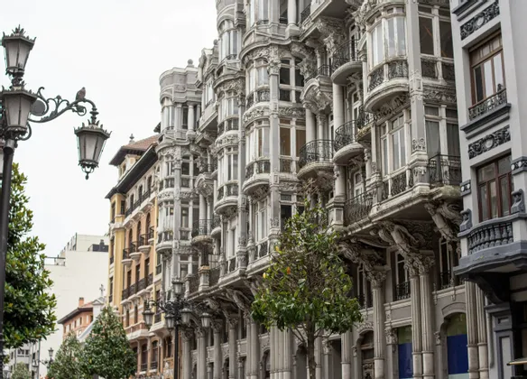 Edifici grigi riccamente decorati con bovindi e balconi dettagliati fiancheggiano una strada cittadina, accanto ad alberi e un lampione d'epoca.