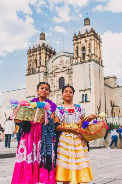 Dos mujeres jóvenes con coloridos vestidos tradicionales sostienen cestas de flores de papel, posando frente a una gran iglesia de piedra.