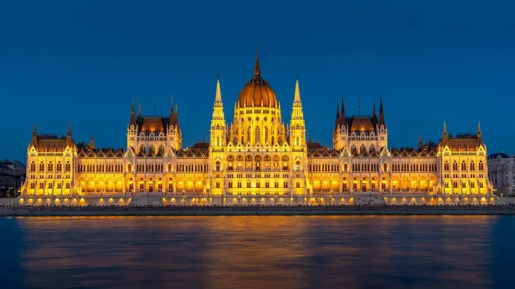 Un grande edificio sontuoso illuminato da luci dorate, che si riflette in un fiume al tramonto sotto un cielo blu intenso.