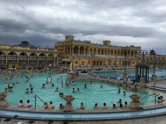Molte persone si rilassano in una grande piscina termale all'aperto di fronte a un imponente edificio giallo sotto un cielo nuvoloso.