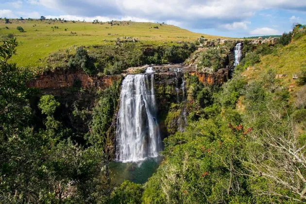 A large waterfall cascades over a rocky cliff into a pool of water, surrounded by dense green trees and rolling hills.
