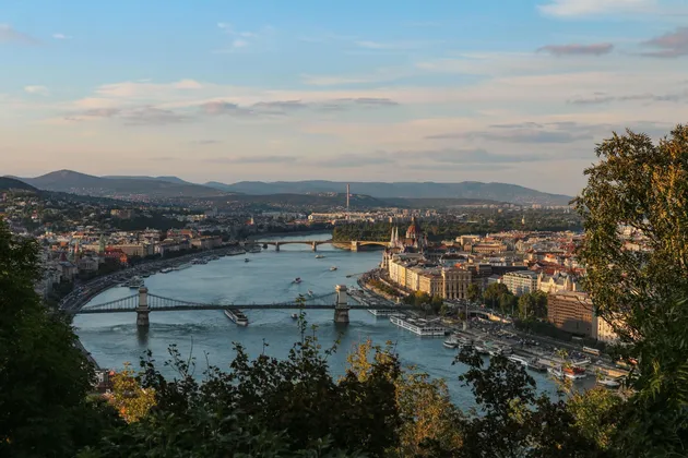 Veduta panoramica di una città con un ampio fiume, diversi ponti e imbarcazioni, con colline distanti sullo sfondo.