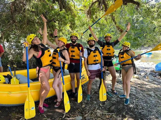 Un viaggio di gruppo WeRoad con caschi e giubbotti di salvataggio in posa con pagaie accanto a un gommone sulla riva di un fiume.