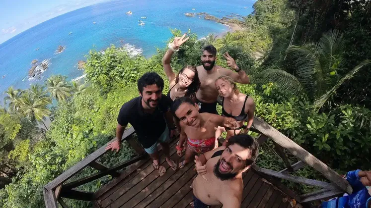 Un gruppo WeRoad scatta un selfie sorridente da un belvedere in legno che si affaccia su una costa tropicale con acque azzurre.