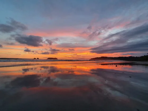 Un tramonto vibrante con nuvole arancioni, rosa e blu che si riflettono sulla sabbia bagnata e specchiata di una spiaggia.